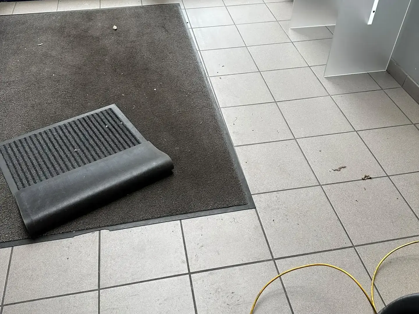 Hall d'entrée avant intervention : sol mat, débris sur le carrelage et paillasson replié.